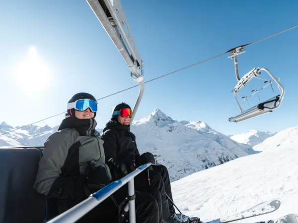 Snowy adventures on winter holidays in the Alps Zwei Skifahrer in Skikleidung auf einem Sessellift mit verschneiten Bergen im Hintergrund