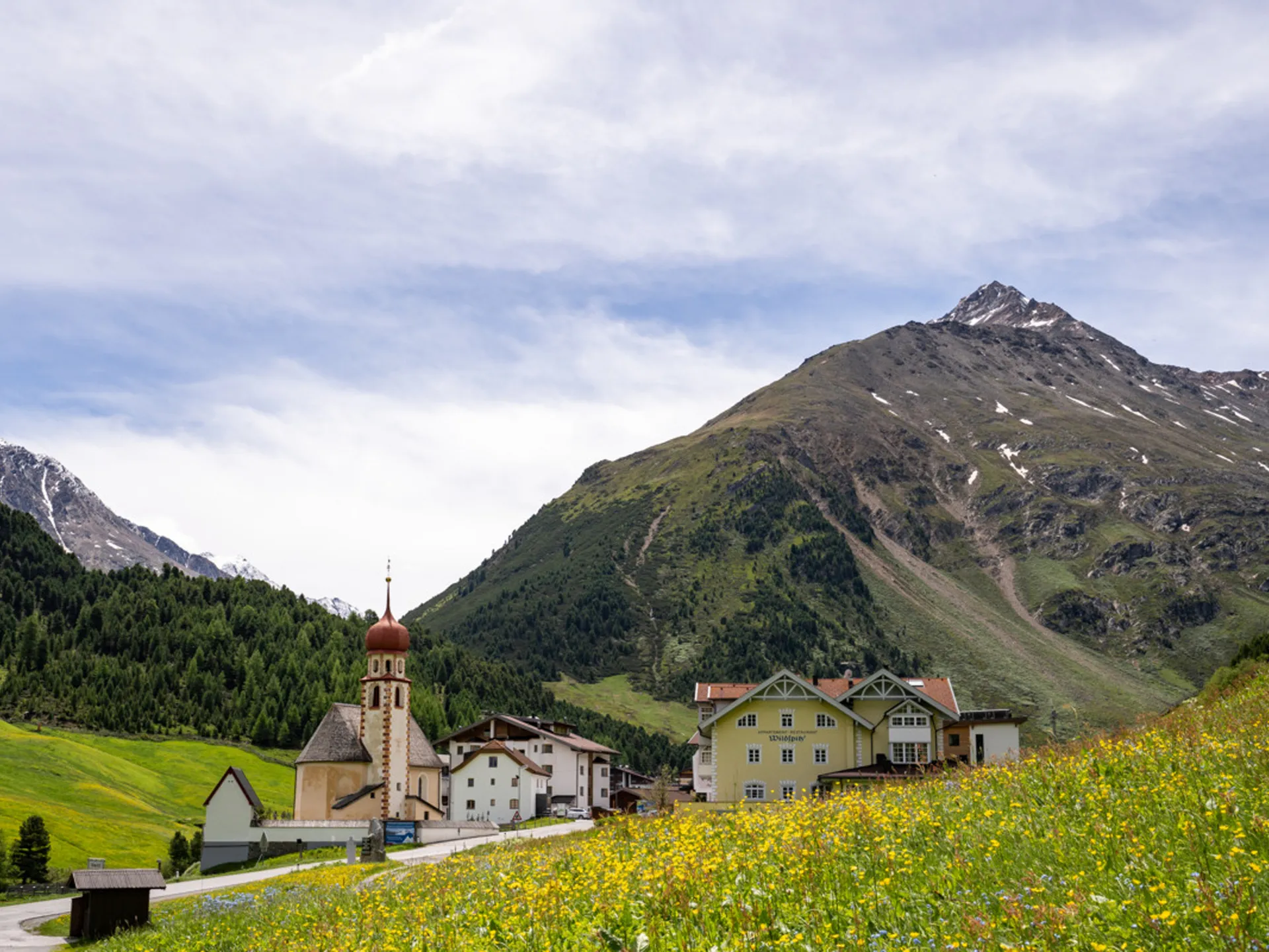 Weisskugel: dein Gasthof in Vent Dorf mit Kirche und Häusern vor Berglandschaft und blühender Wiese